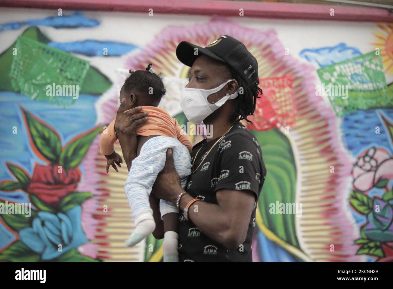 A Haitian migrant carries his daughter in front of a mural of the Virgin of Guadalupe inside the Casa de Acogida Formación y Empoderamiento de la Mujer Migrante y Refugiada (CAFEMIN), located in Mexico City, Mexico, on September 27, 2021, which offers lodging and food to migrants who recently went to various refugee support offices to try to complete a procedure that would allow them to stay longer in Mexico given the difficulties their compatriots are having in entering the United States during the COVID-19 health emergency. (Photo by Gerardo Vieyra/NurPhoto) Stock Photo