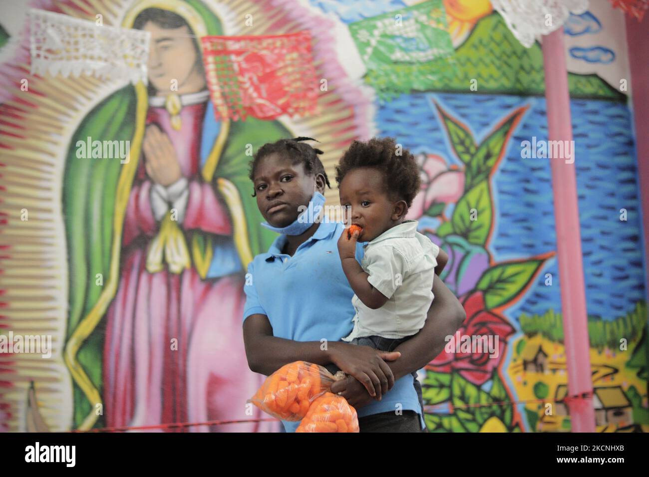 A Haitian migrant carries her daughter in front of a mural of the Virgin of Guadalupe inside the Casa de Acogida Formación y Empoderamiento de la Mujer Migrante y Refugiada (CAFEMIN), located in Mexico City, Mexico, on September 27, 2021, which offers accommodation and food to migrants who recently went to various refugee support offices to try to obtain a permit to stay longer in Mexico in view of the difficulties their compatriots are having in entering the United States during the COVID-19 health emergency. (Photo by Gerardo Vieyra/NurPhoto) Stock Photo