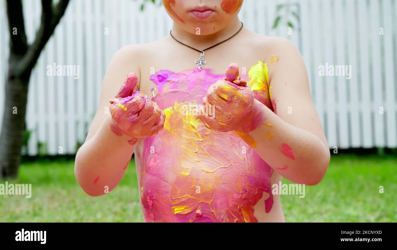 a little child, a four-year-old boy playing, painting with finger paints, decorating himself, in the garden, sitting on a blanket, on grass, lawn, in the summer. he's having fun. High quality photo Stock Photo