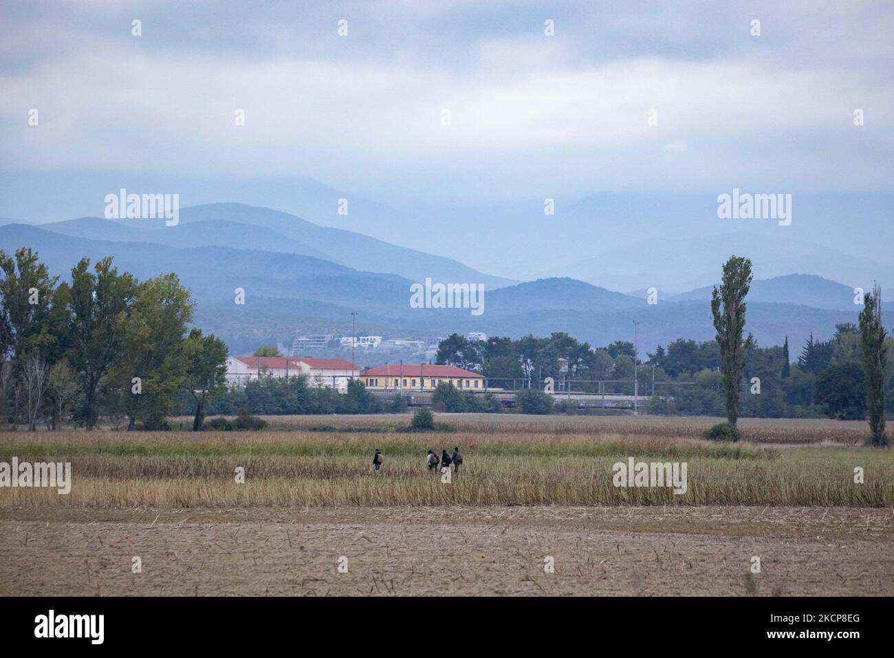 Migrants are seen walking in the fields with the iconic yellow train station of Idomeni and houses in N. Macedonia in the background. Asylum Seekers are trying to cross the Greek-North Macedonian borders to follow the Balkan Route, famous in 2015-2016 during the Syrian refugee crisis, from Idomeni, Greece to Gevgelija, North Macedonia following the train rails and railway station and then reach central and northern Europe. Refugees and Migrants are seen walking in the fields in the Greek side, before the fence that separates the two countries, trying to reach and pass the borders. The small gr Stock Photo