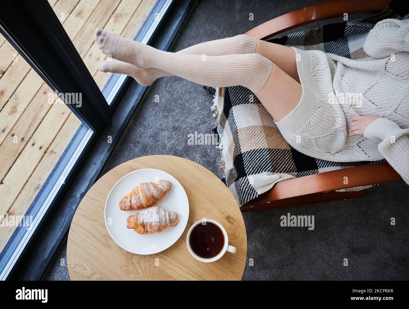 Place with croissants and cup of tea on the table. Cropped view of young woman's legs near window inside contemporary barn house. Stock Photo