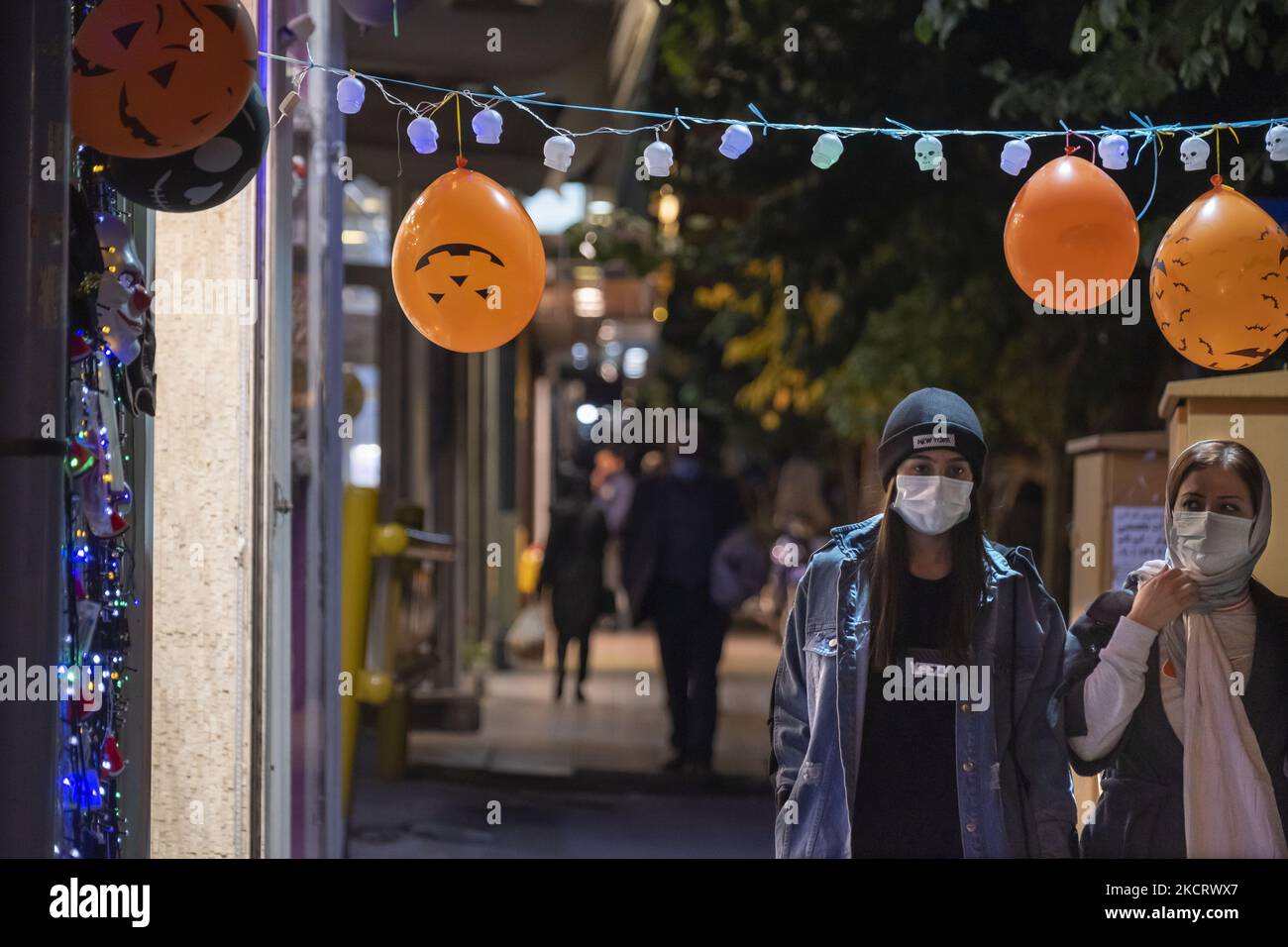 Two Iranian women wearing protective face masks walks past halloween products that are hanged out of a toy shop in downtown Tehran, October 30, 2021. (Photo by Morteza Nikoubazl/NurPhoto) Stock Photo