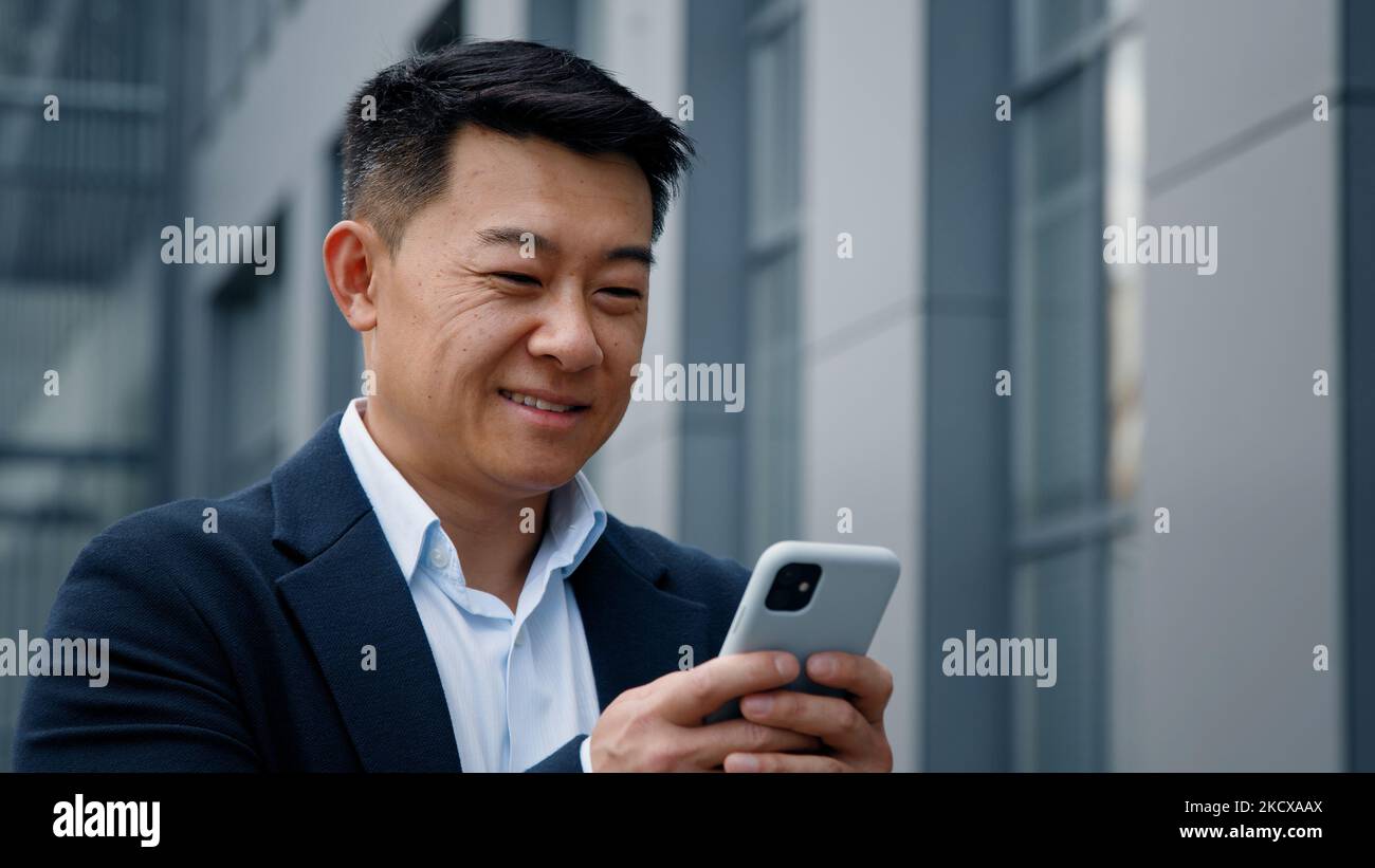 Close up smiling laughing Asian middle-aged Korean businessman man employer entrepreneur laugh looking at mobile phone chatting browsing smile Stock Photo