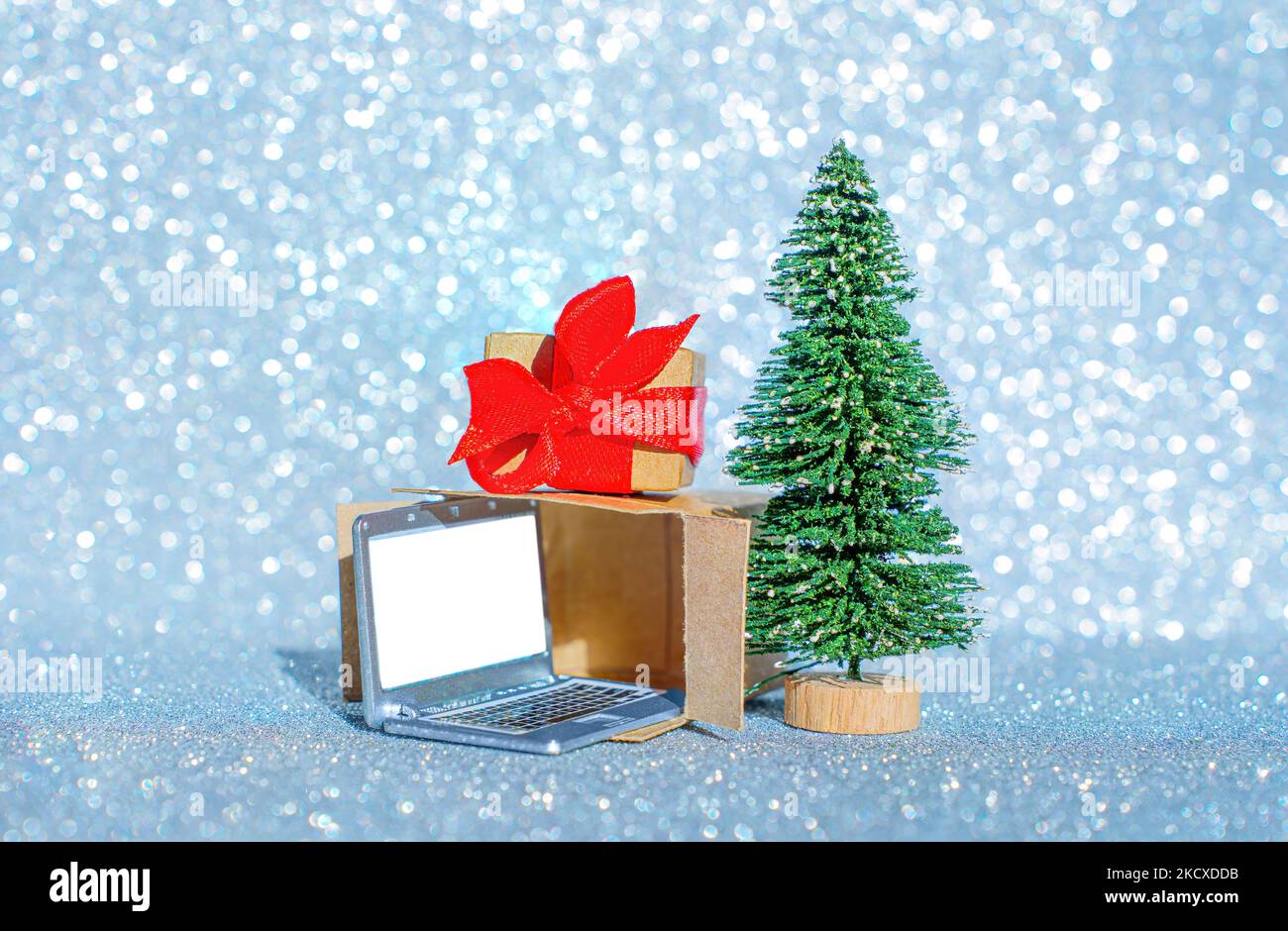 Unwrapped gift box with a miniature laptop by a toy christmas tree with glittering background