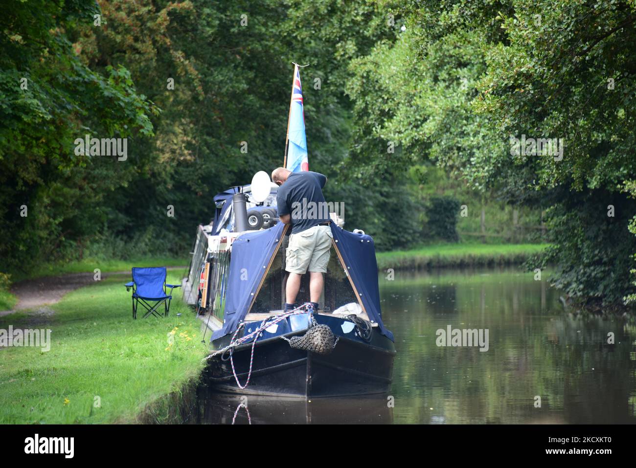 A man stands on the bow of his narrowboat, seemingly repairing it whilst it is moored on a summer afternoon. Stock Photo