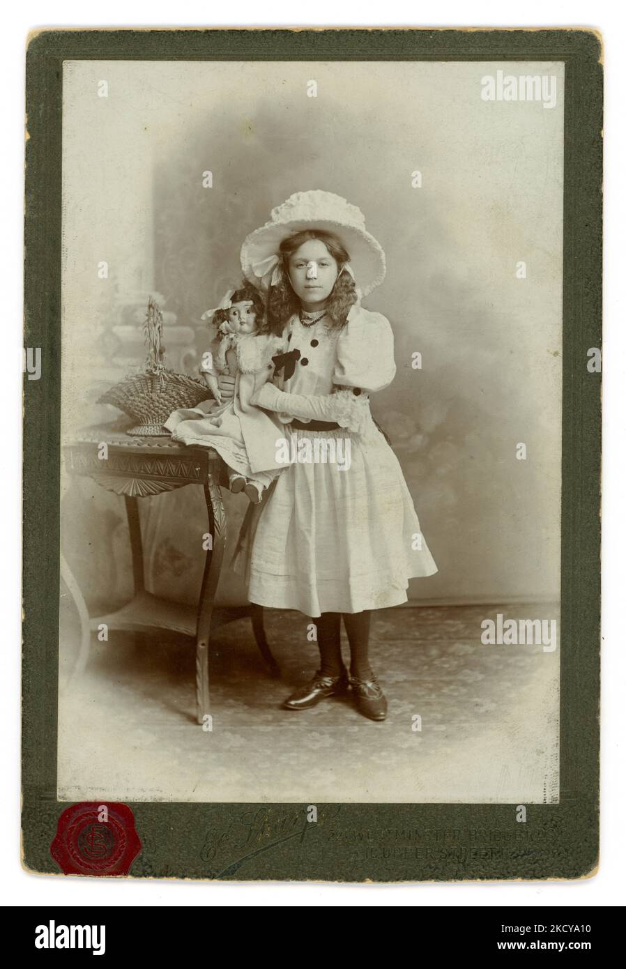 Edwardian era studio portrait cabinet card of pretty looking girl from a privileged, upper / middle class family wearing a large white hat, beautiful white dress (party ?)  with puffed sleeves, white gloves and tights and holding her china doll. Perhaps a birthday photograph. On reverse is written Clara R. J. Whant, aged 11 1909. From the studio of Edward Sharp Islington and Westminster Bridge London, U.K. Dated 1909. Stock Photo