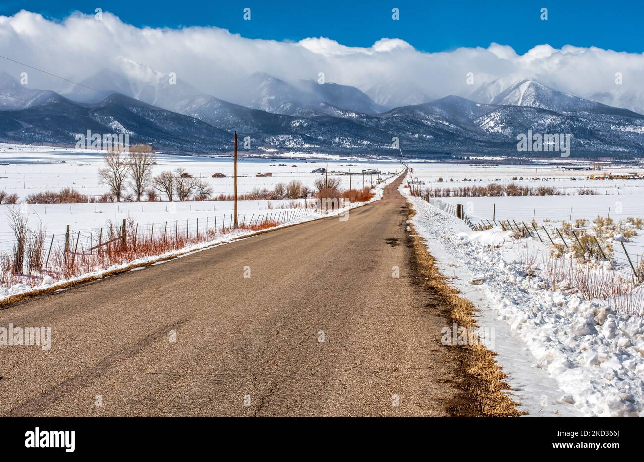 A rural country road through snow covered ranchland leads to the even more snow covered mountains of the Sangre de Cristo range in the Colorado Rocky Stock Photo