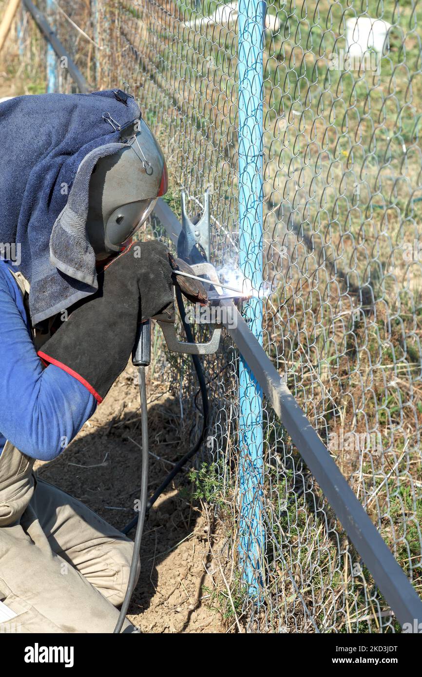 Male welder in brown overalls, protective mask and gloves welds metal pipes to vertical bases for further attachment of metal sheets using electric ar Stock Photo