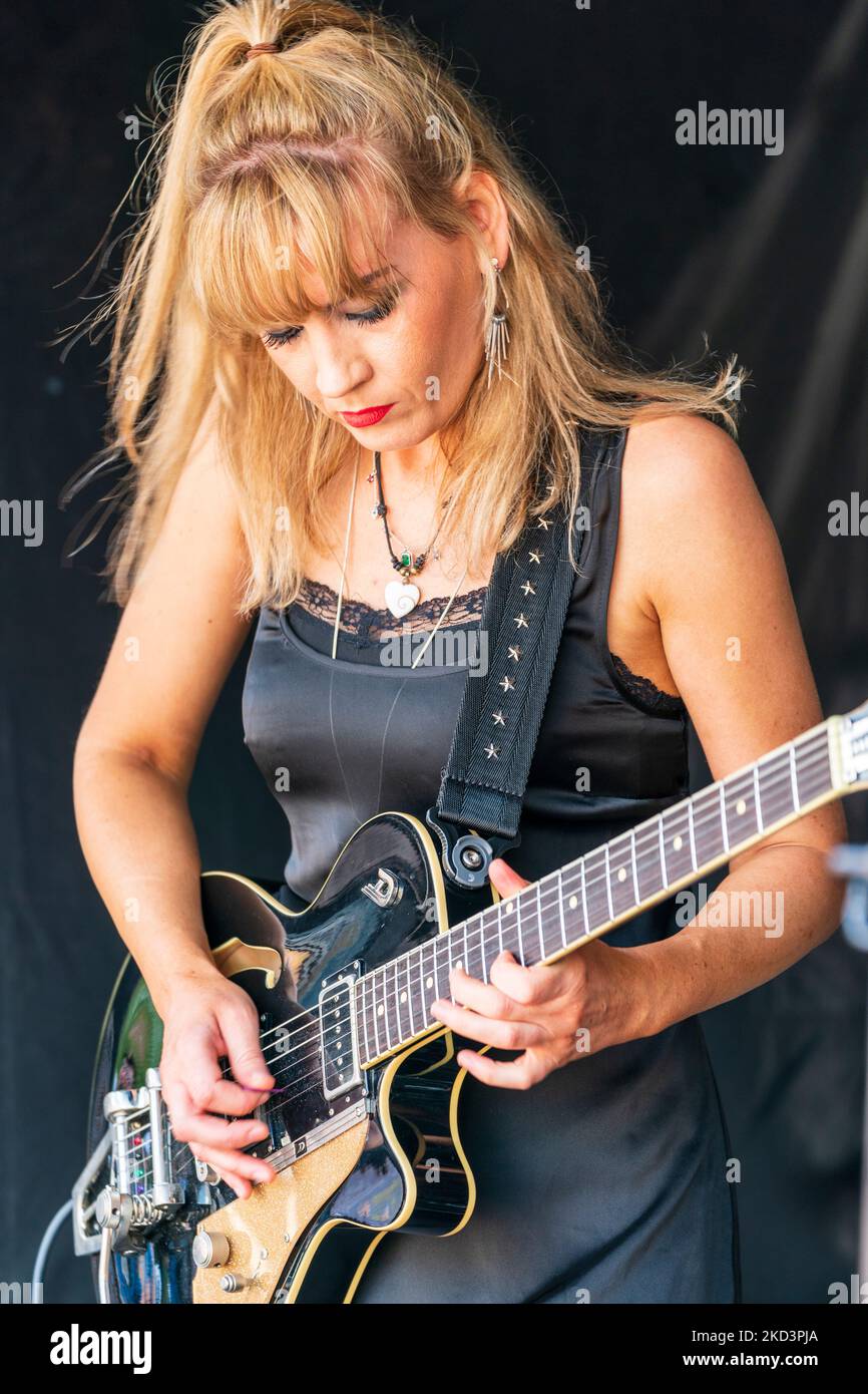 Close up of blonde woman playing electric guitar. Head bowed down as she concentrates on the cords. Black curtain background. Stock Photo