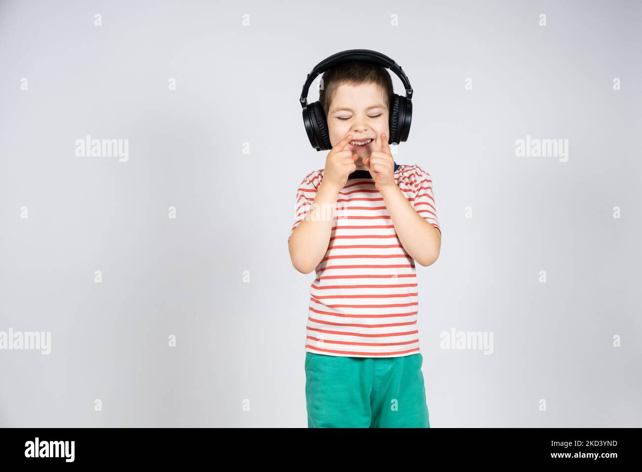 Cute 5 year old boy in big black headphones laughs and points his fingers up on a white background, a place for text to copy space Stock Photo