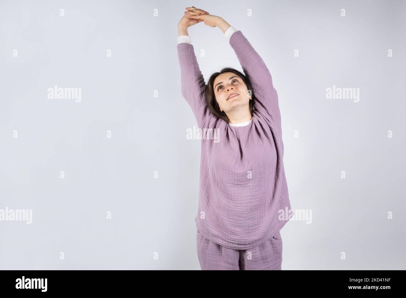 Young brunette woman in muslin lavender pajamas sleep clothes stretches by raising her arms up. Stock Photo