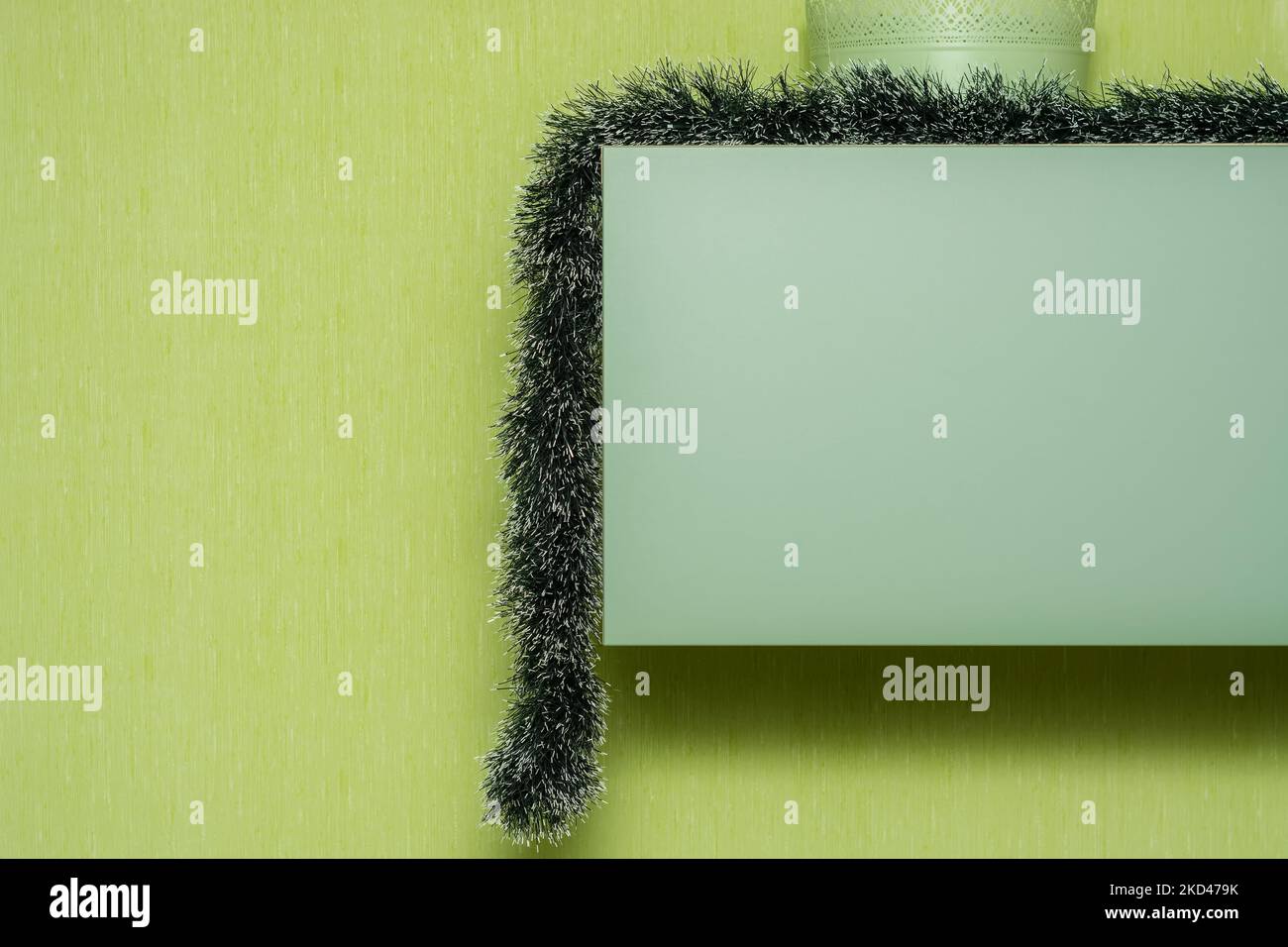 Long green Christmas tree garland hangs from a wall cabinet against a green wall. Stock Photo
