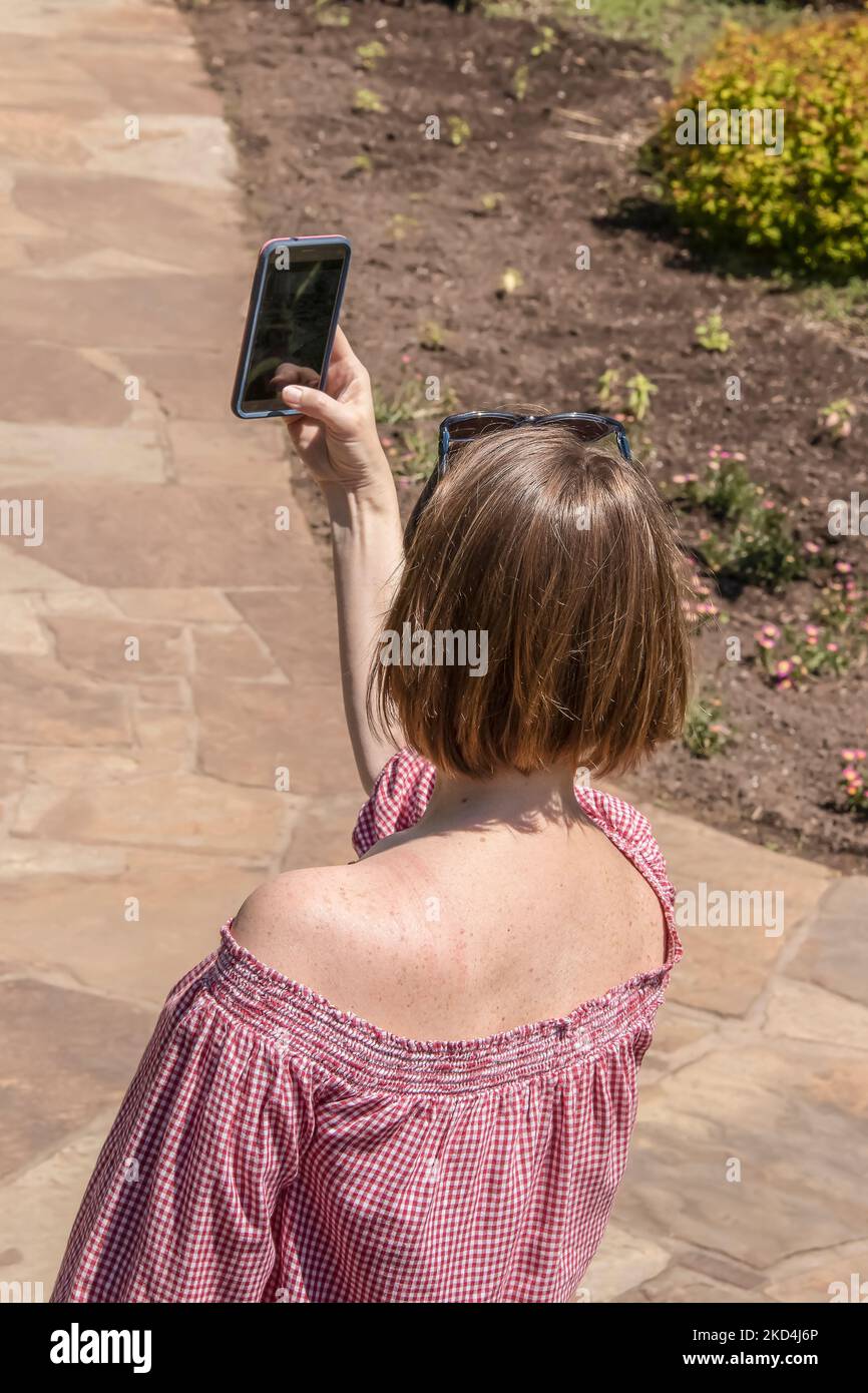 Woman with short bob and sunglasses on her head and off-shoulder blouse takes a selfie in a garden - back view Stock Photo
