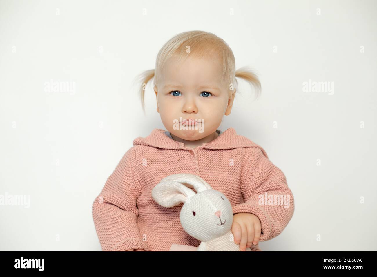 Little baby girl play with stuffed animal isolated on white. Toddler with sad facial expression holding teddy bunny. Blonde haired child in pink Stock Photo