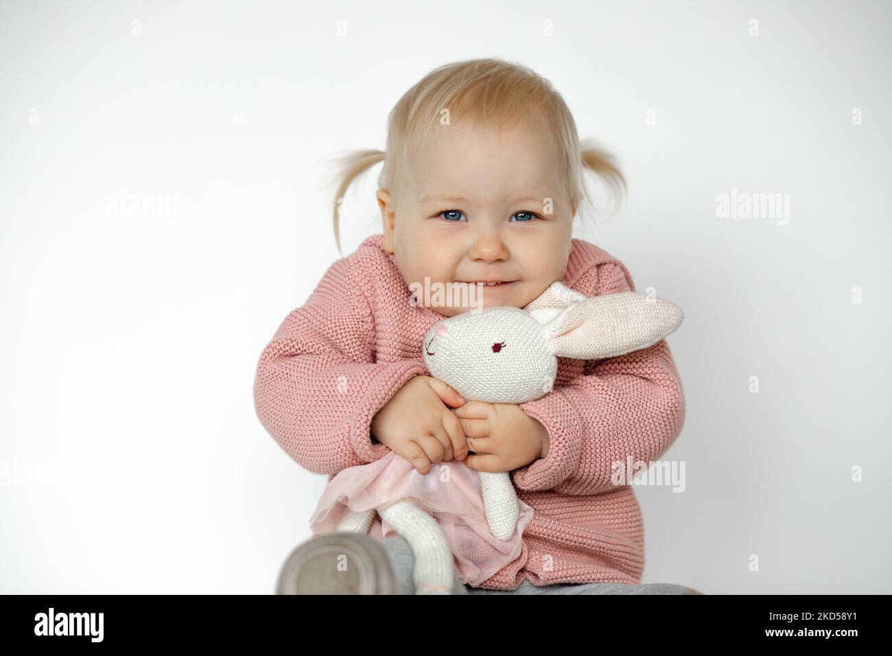 Lovely baby girl play with stuffed animal, isolated on white. Overjoyed toddler gladly embracing teddy bunny. Blonde haired child in pink clothes Stock Photo
