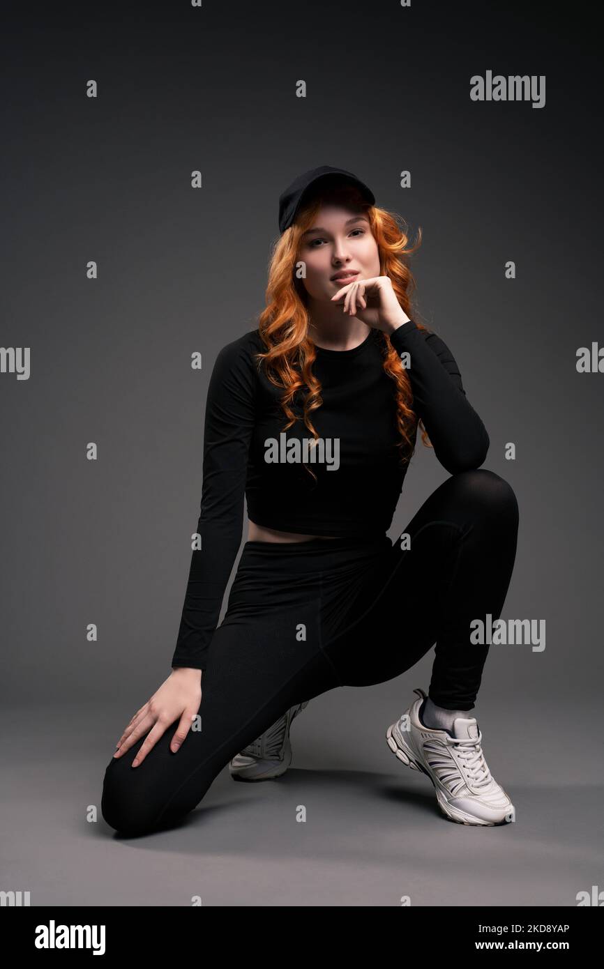 Stylish redhead lady adjusting cap and smiling at camera in studio Stock Photo