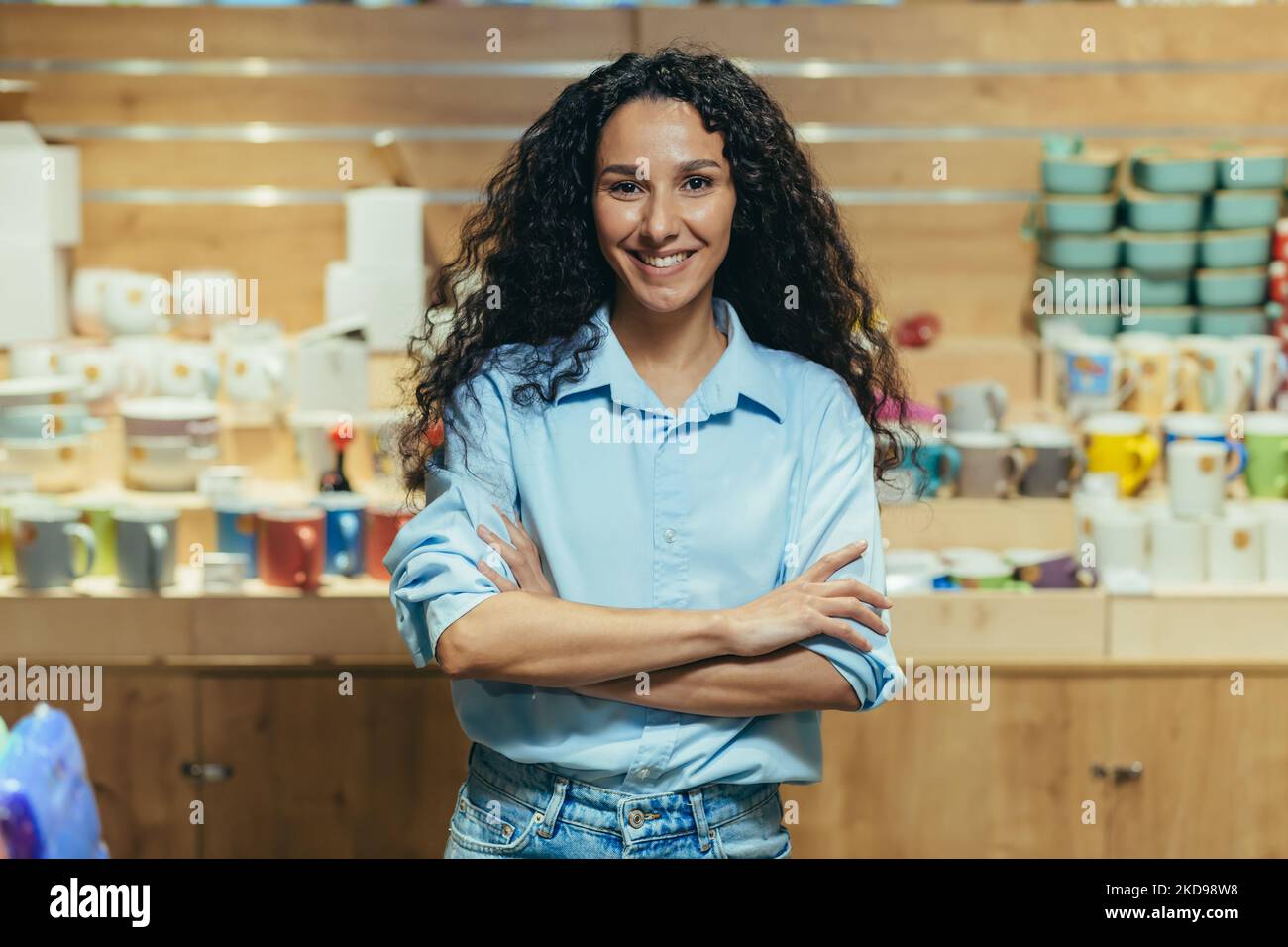 Portrait of business owner, hispanic woman managing gift shop, woman with curly hair looking at camera and smiling with arms crossed. Stock Photo