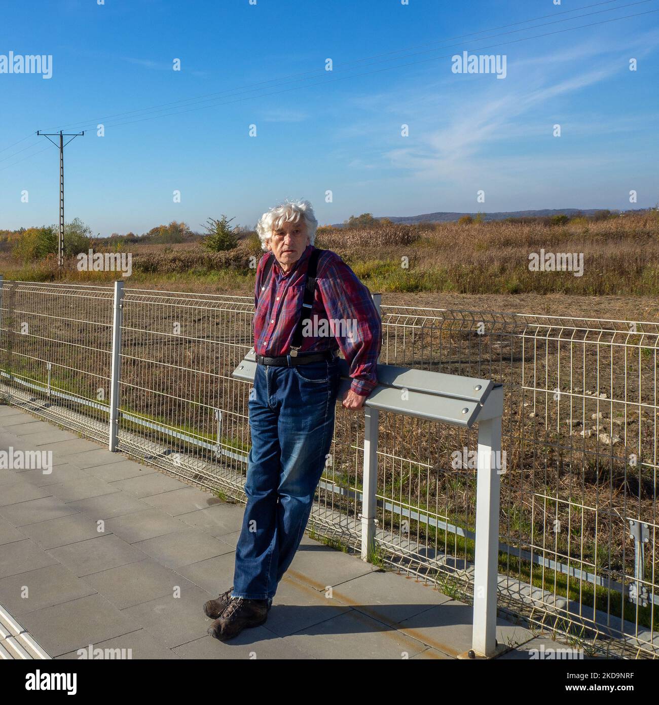 Elderly gray-haired man in blue jeans leaning  against a wooden modern leaning bench or leaning stand or lean bar (no established name yet) waiting fo Stock Photo