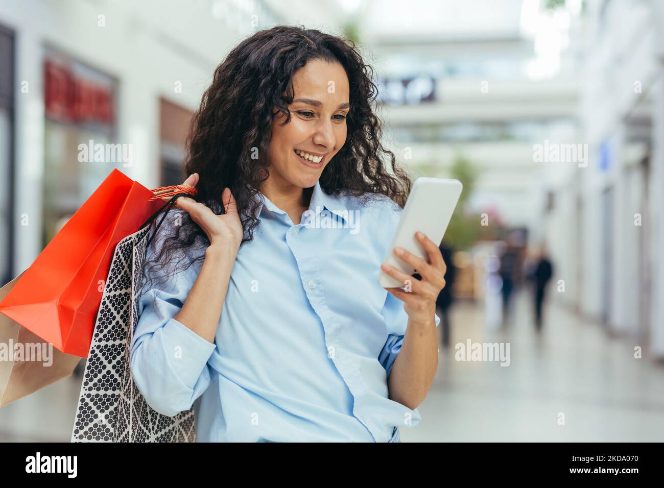 Happy and successful woman shopping for clothes in a supermarket store, Hispanic woman holding a smartphone reading online messages and browsing offers with discounts and sales Stock Photo