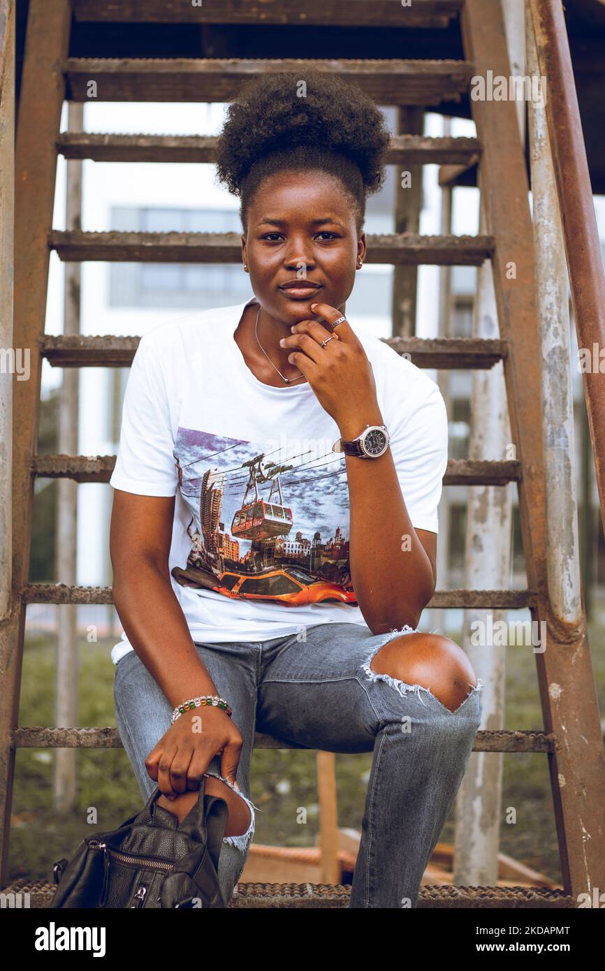 A vertical of a black lady with afro hair and in a white shirt sitting on a staircase. Stock Photo