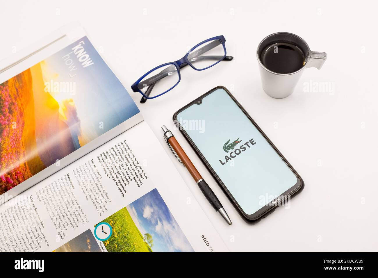 In this photo illustration a Lacoste logo seen displayed on a smartphone screen on a desk next to a cafe, a pen, glasses and a magazine in Athens, Greece on June 27, 2022. (Photo by Nikolas Kokovlis/NurPhoto) Stock Photo