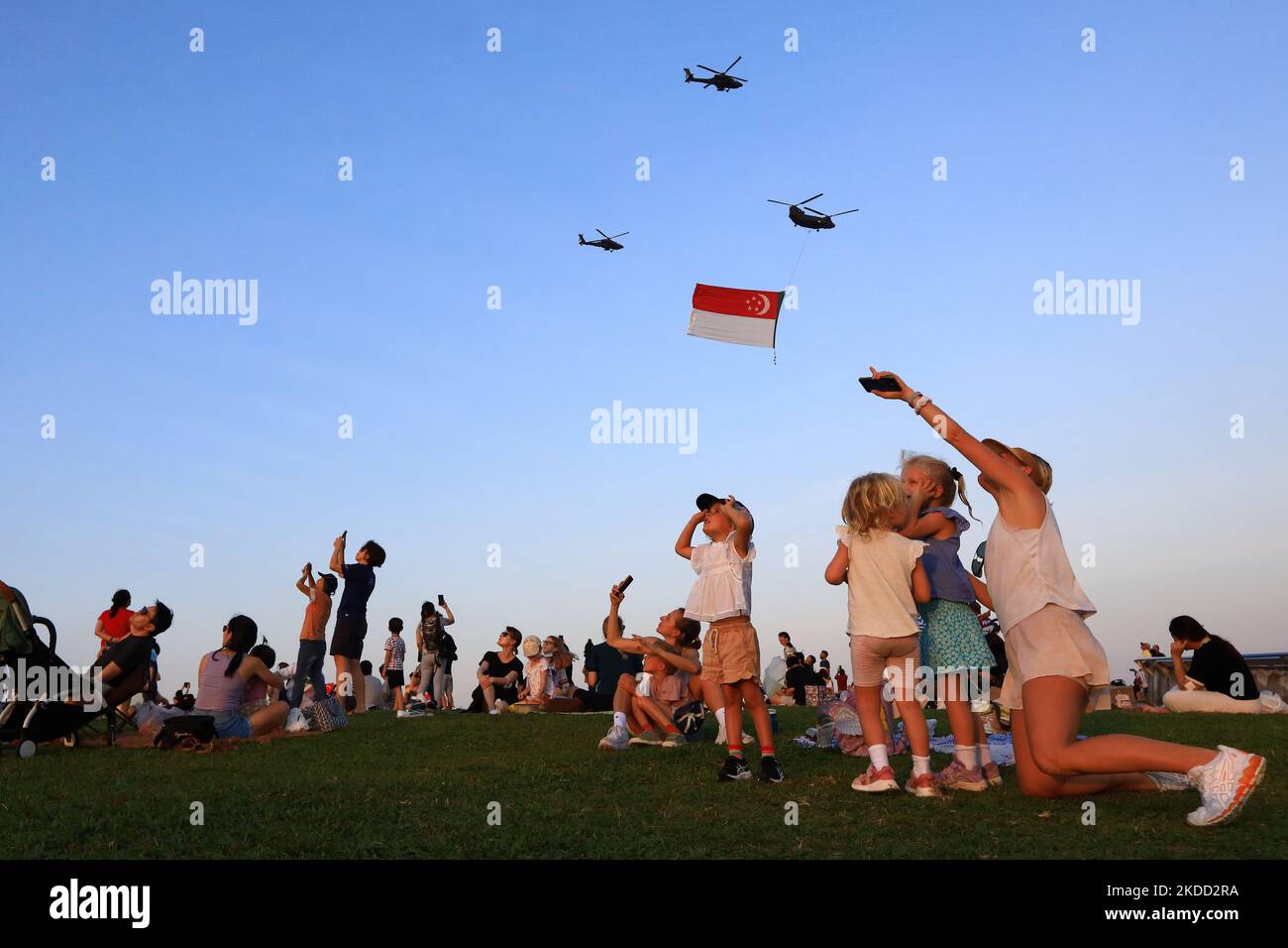 People react and take photos as the CH-47 Chinook helicopter, escorted by two AH-64D Apache helicopters make a flypast of the State Flag during the National Day Parade rehearsal at Marina Bay on July 2, 2022 in Singapore. (Photo by Suhaimi Abdullah/NurPhoto) Stock Photo