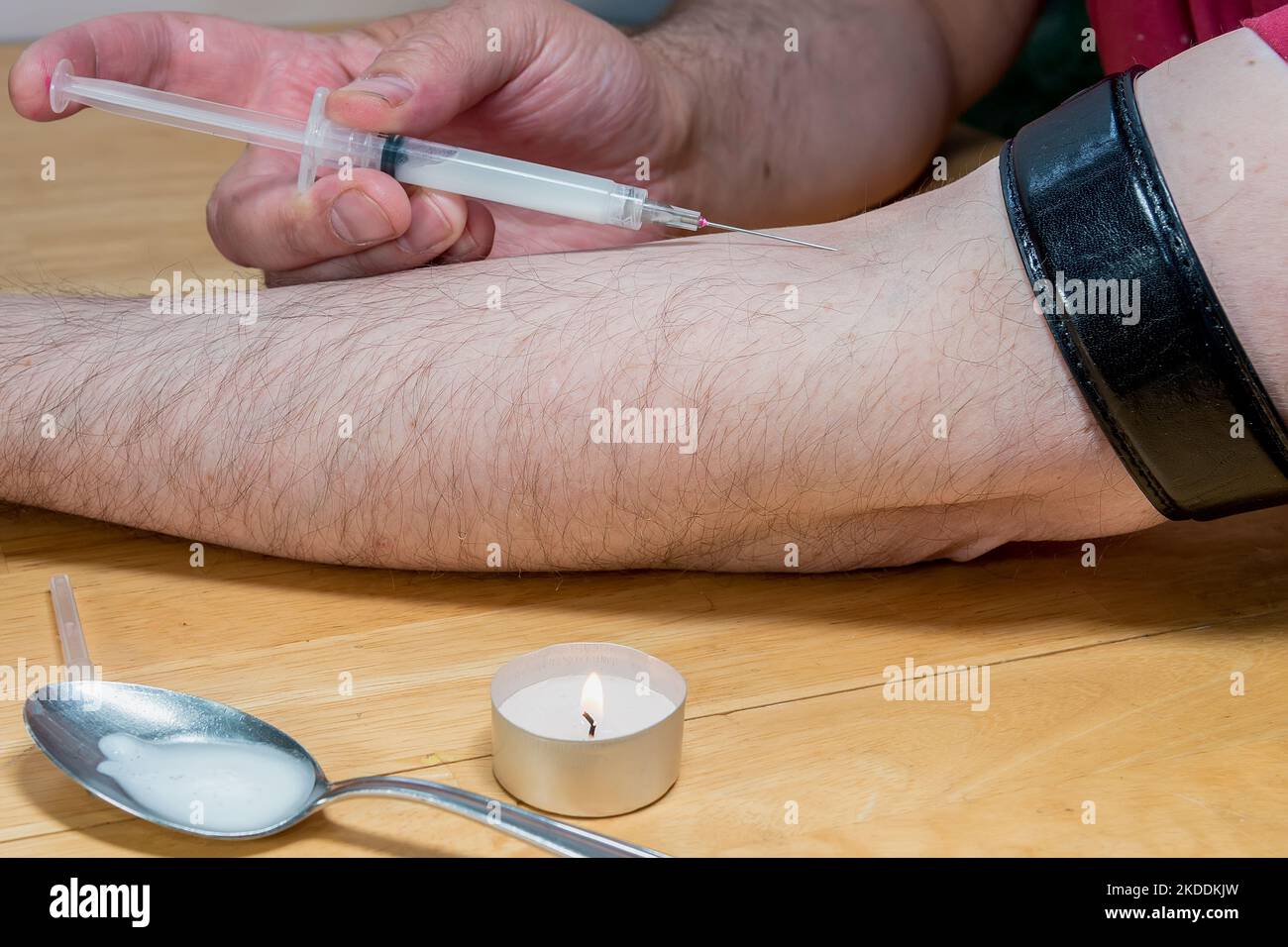 Junkie injecting drugs into his arm. A belt is wrapped around his upper arm. Candle and spoon with partially dissolved drugs in foreground. Stock Photo