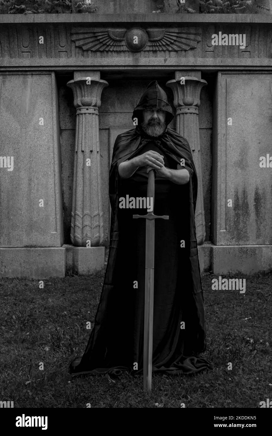 Man with a beard wearing a black hooded cape. He is a knight holding a large sword on the ground. Grave crypt in the background. Black and white. Stock Photo