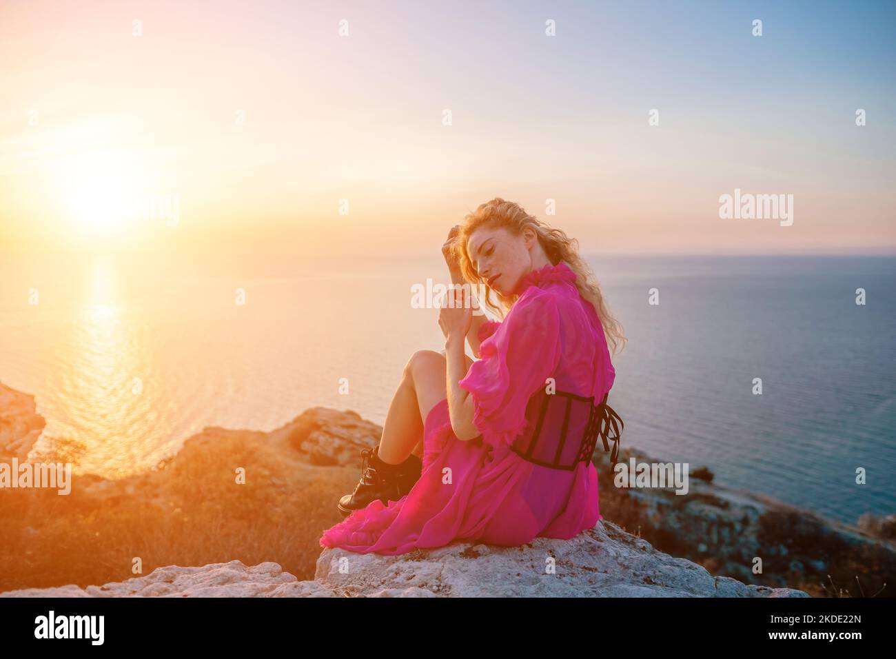 Beautiful young caucasian woman with curly blond hair and freckles. Cute redhead woman portrait in a pink long dress posing on a volcanic rock high Stock Photo