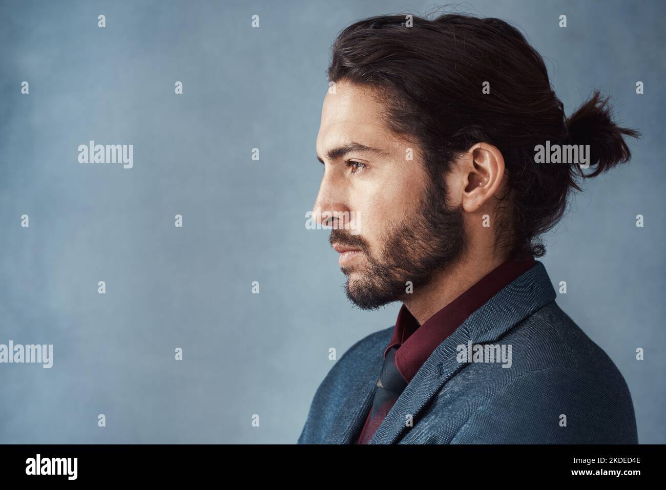 He knows how to wear a man bun. Studio shot of a handsome young man looking thoughtful against a grey background. Stock Photo