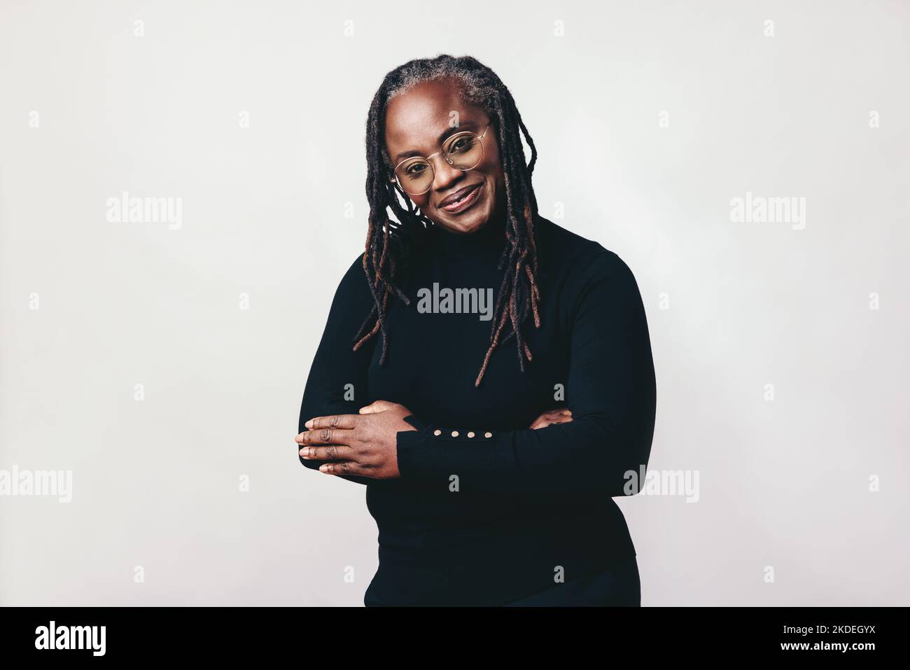Professional woman with dreadlocks smiling at the camera while wearing eyeglasses. Happy mature woman standing against a grey background with her arms Stock Photo