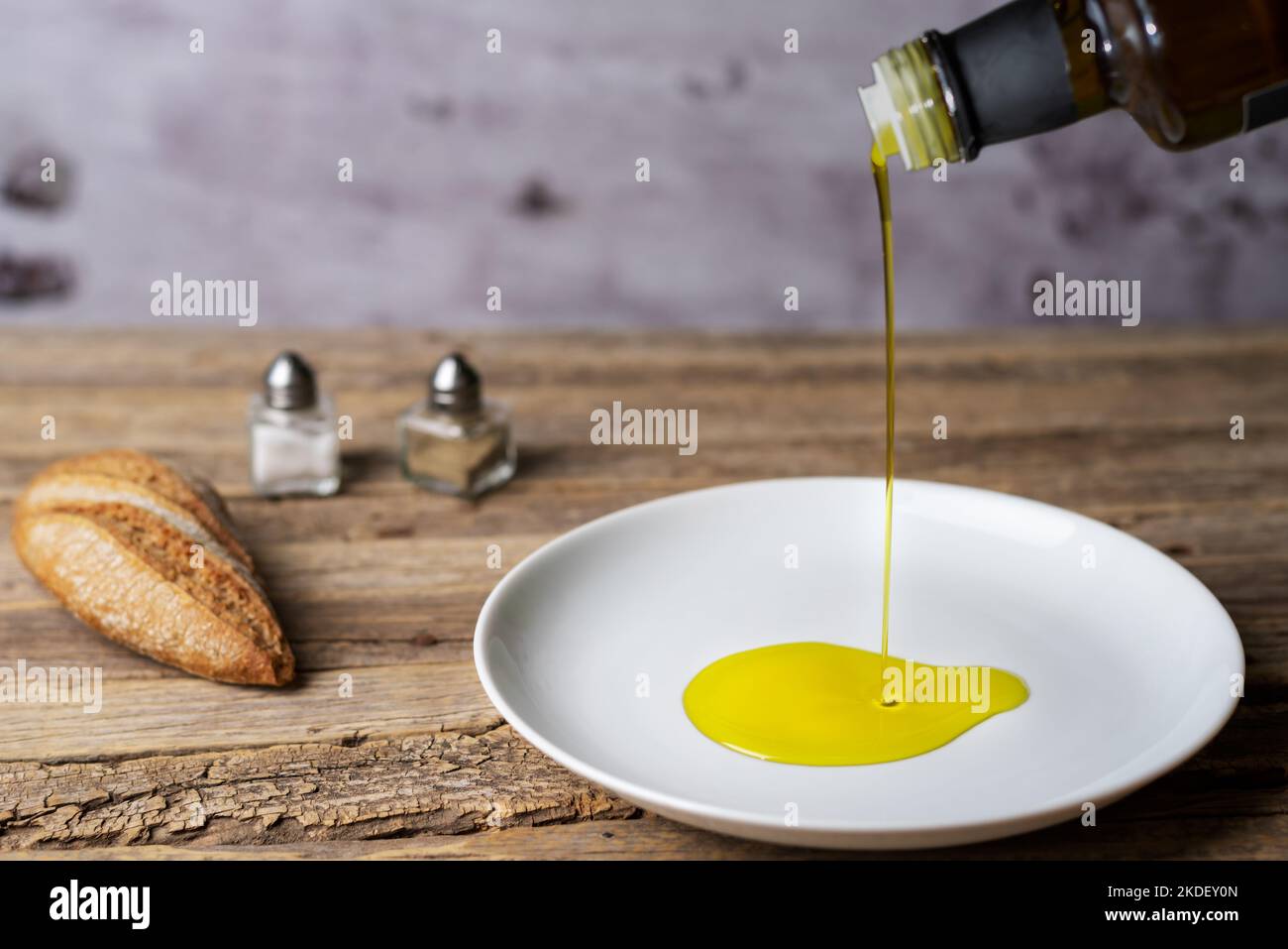 Extra virgin olive oil pouring from a bottle to a round white plate, on a rustic wooden table. Stock Photo