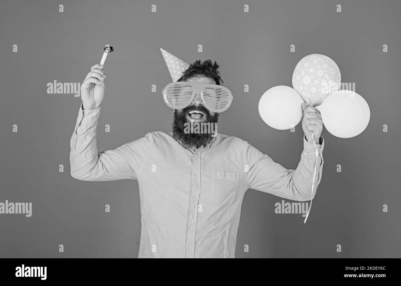 Hipster in giant sunglasses celebrating birthday. Man with beard and mustache on happy face holds party horn, blue background. Celebration concept Stock Photo