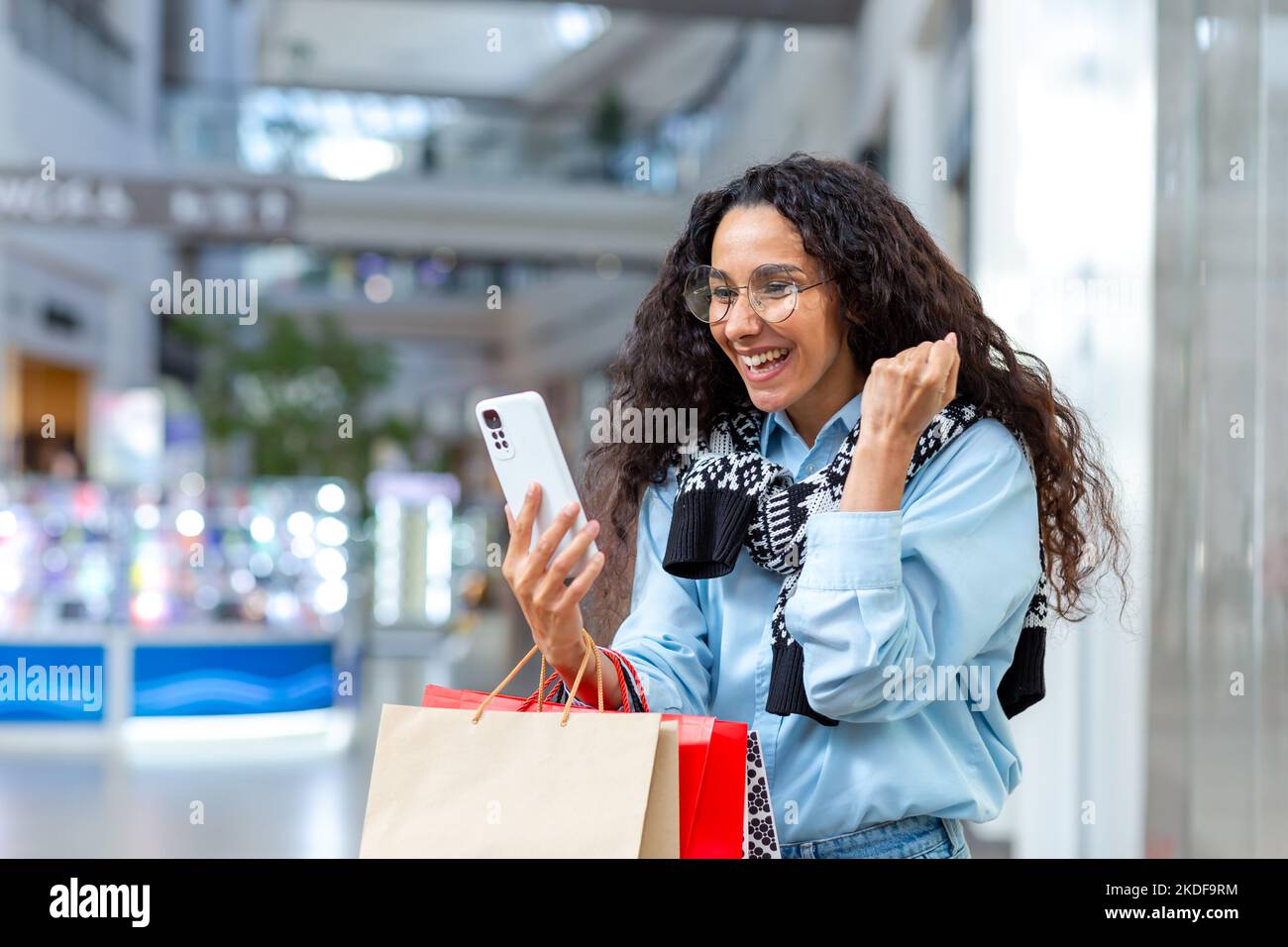 Portrait of happy woman shopper, Hispanic woman inside mall, uses smartphone, browses online discounts and sales, holds hand up, celebrates received offer, win. Stock Photo