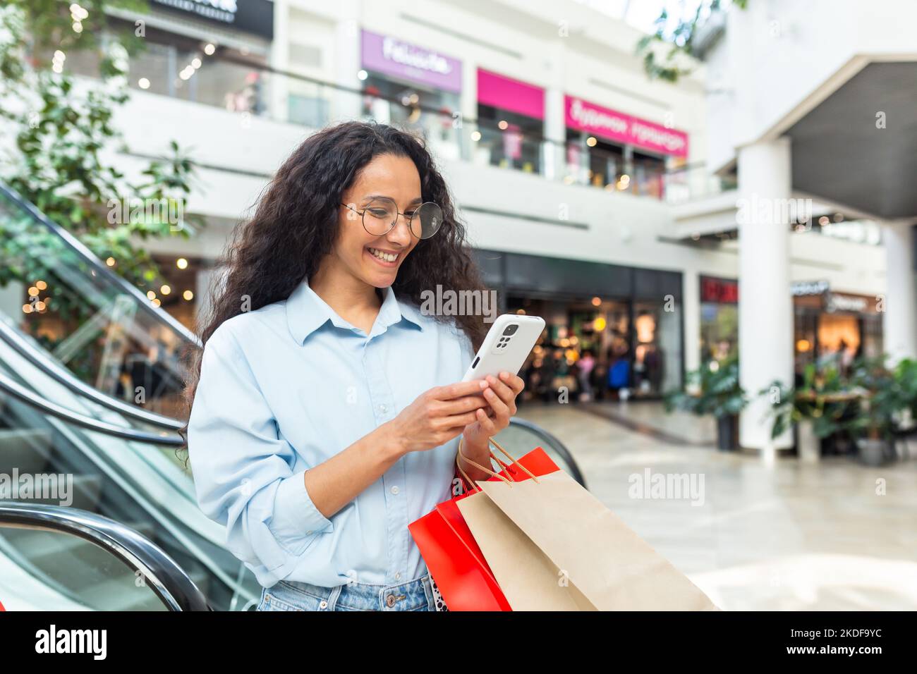 Happy woman shopping for clothes during the period of discounts and promotional offers, Hispanic woman smiling and happy browsing online discounts using smartphone, inside a modern large store. Stock Photo