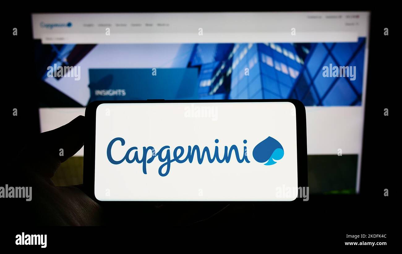 Person holding mobile phone with logo of information technology company Capgemini SE on screen in front of web page. Focus on phone display. Stock Photo