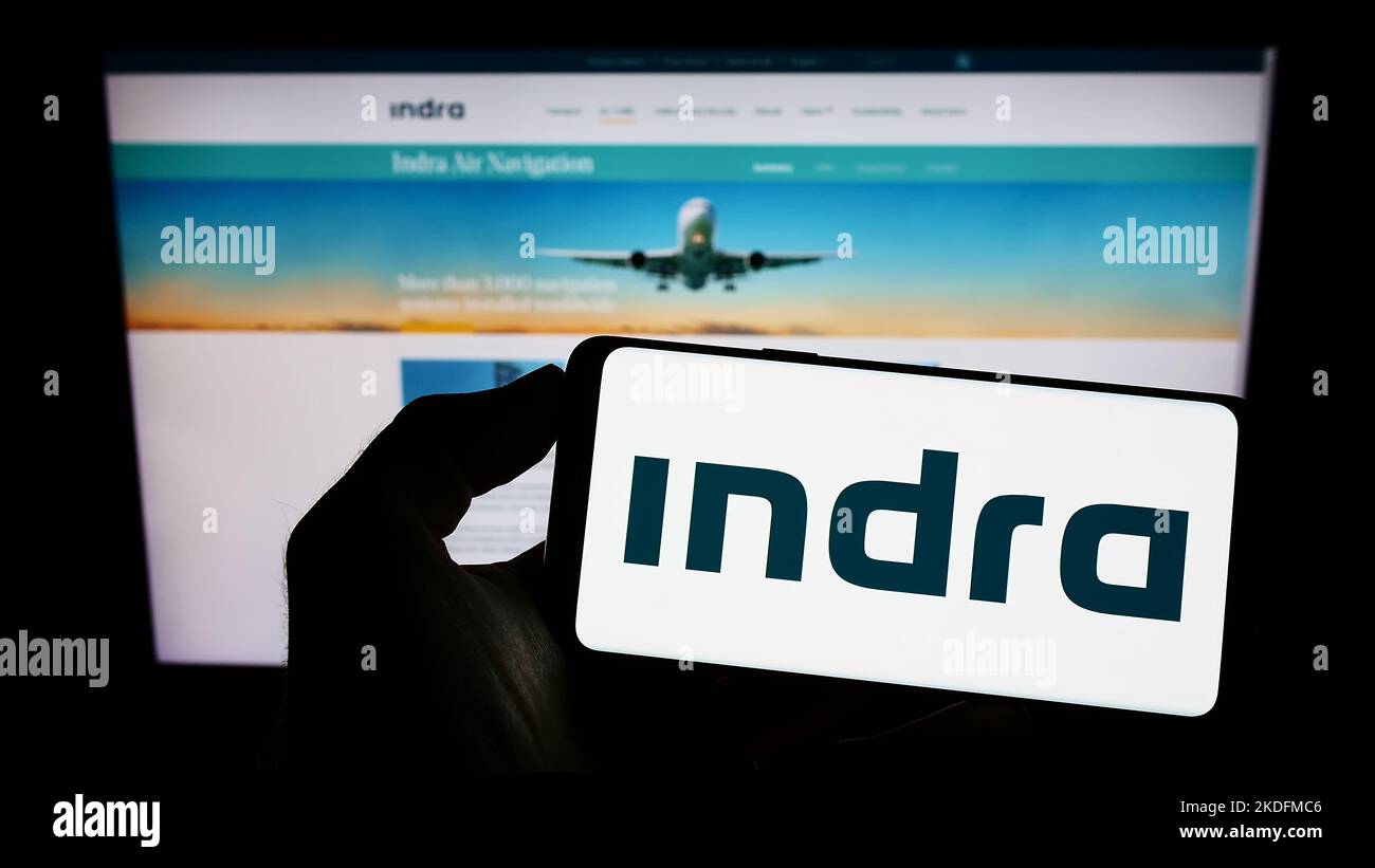Person holding mobile phone with logo of Spanish company Indra Sistemas S.A. on screen in front of business web page. Focus on phone display. Stock Photo