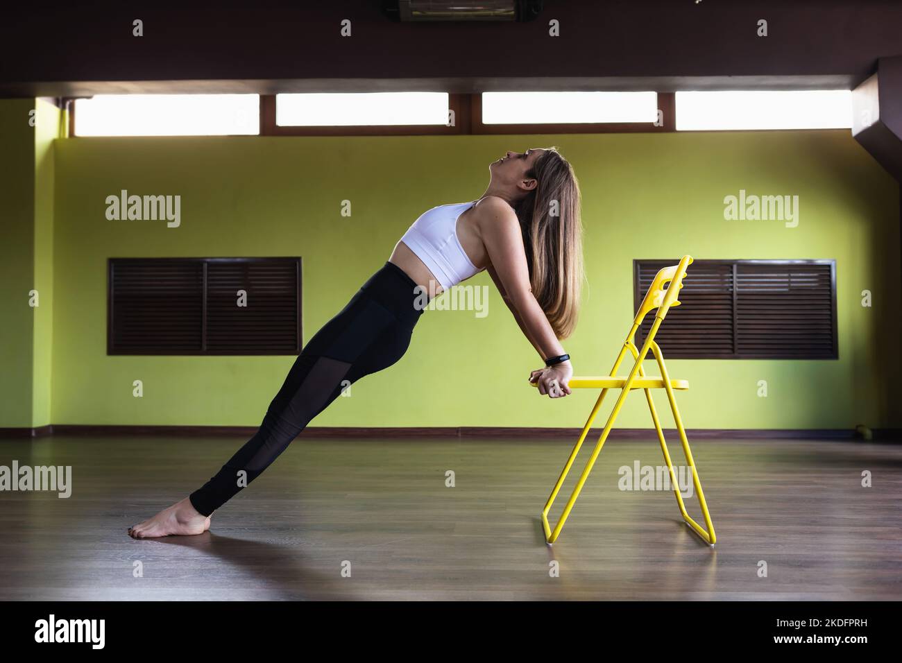 A woman performs an exercise to strengthen the muscles of the back and arms using a chair, doing yoga therapy in the studio, exercising in sportswear Stock Photo