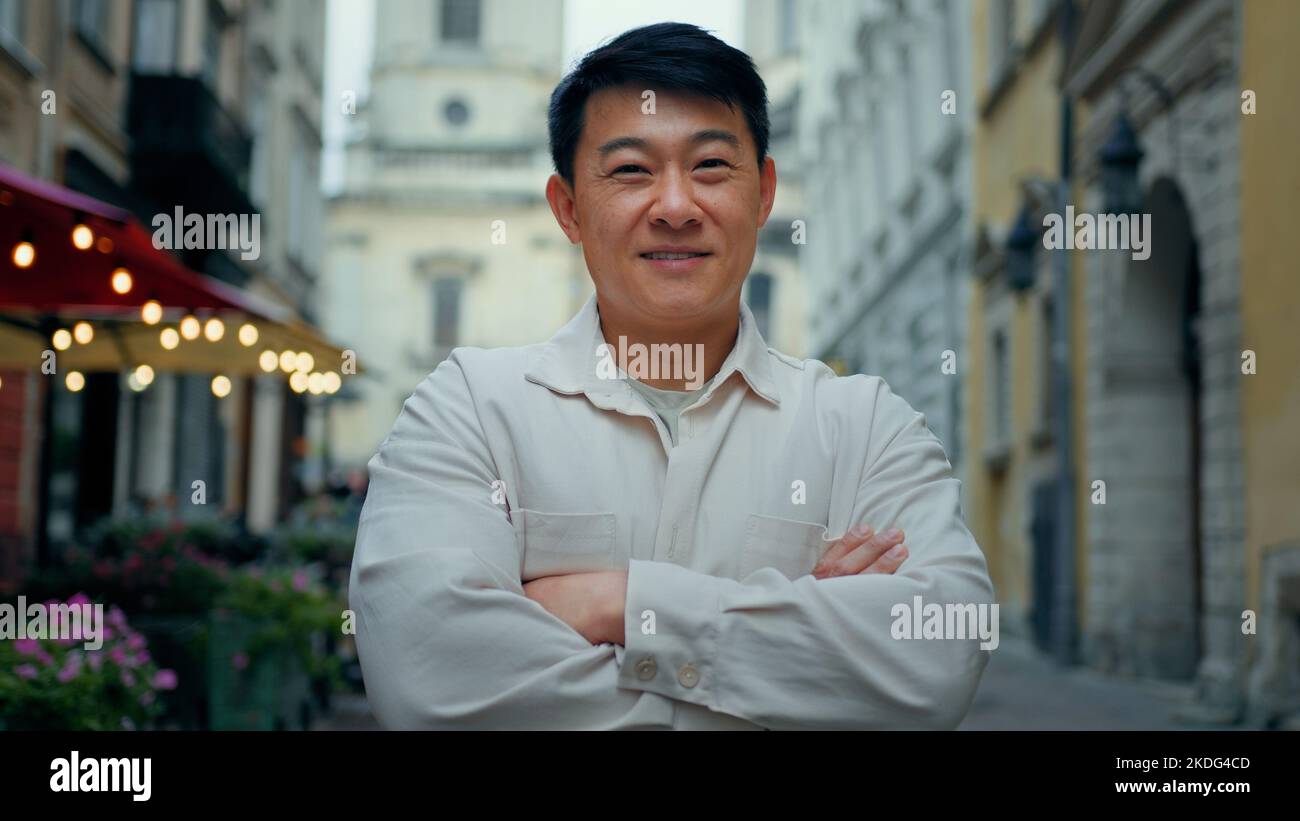 Happy pleased confident man standing outdoor joyful male tourist stands in city center looking at camera smiling asian model posing with crossed arms Stock Photo