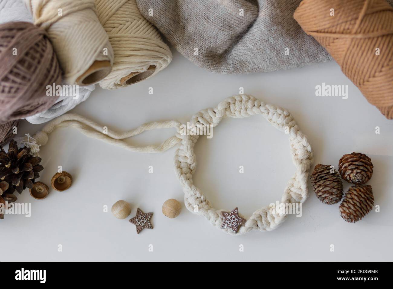 Winter table decor. White macrame wreath, cones, balls of thread and gray knitted sweater on white table. Stock Photo
