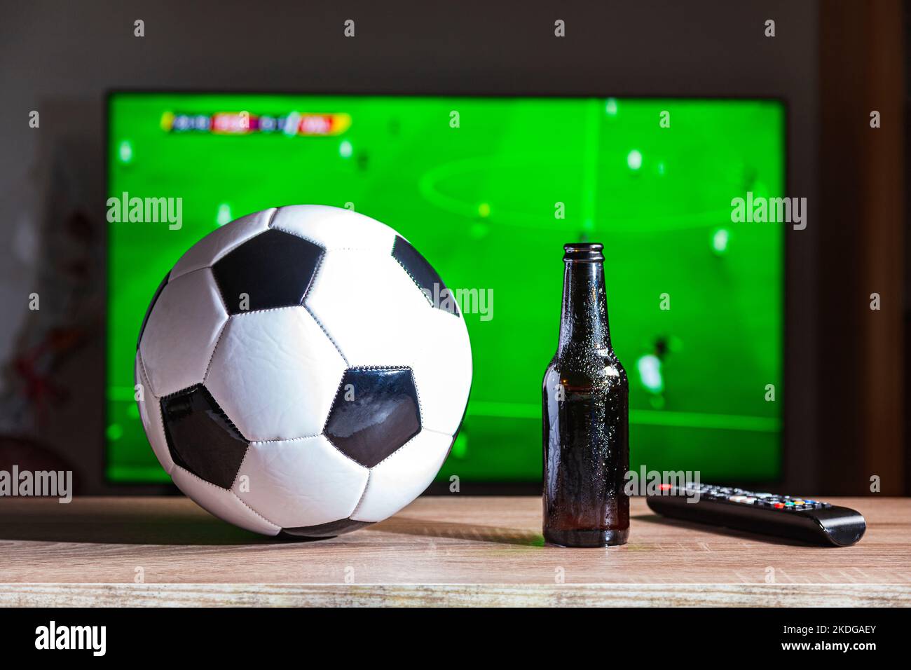 On a table is a soccer ball, a bottle of beer and a remote control. In the background, out of focus, there is a television set on which a football gam Stock Photo