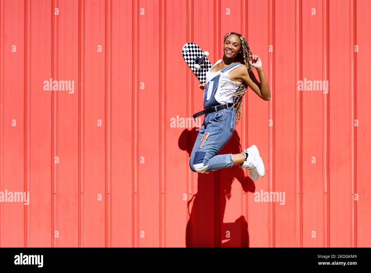 Black woman dressed casual, wtih a skateboard jumping with happiness on red urban wall background. Stock Photo