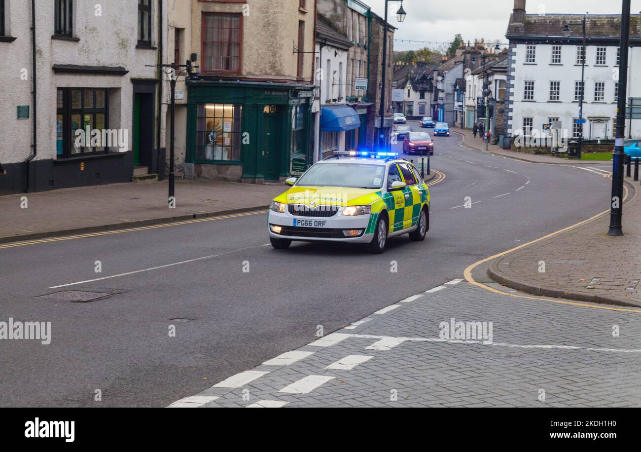 An ambulance car attending an emergency incident speeding through the main street in Kendal, Cumbria, with blue flashing lights and headlights on Stock Photo