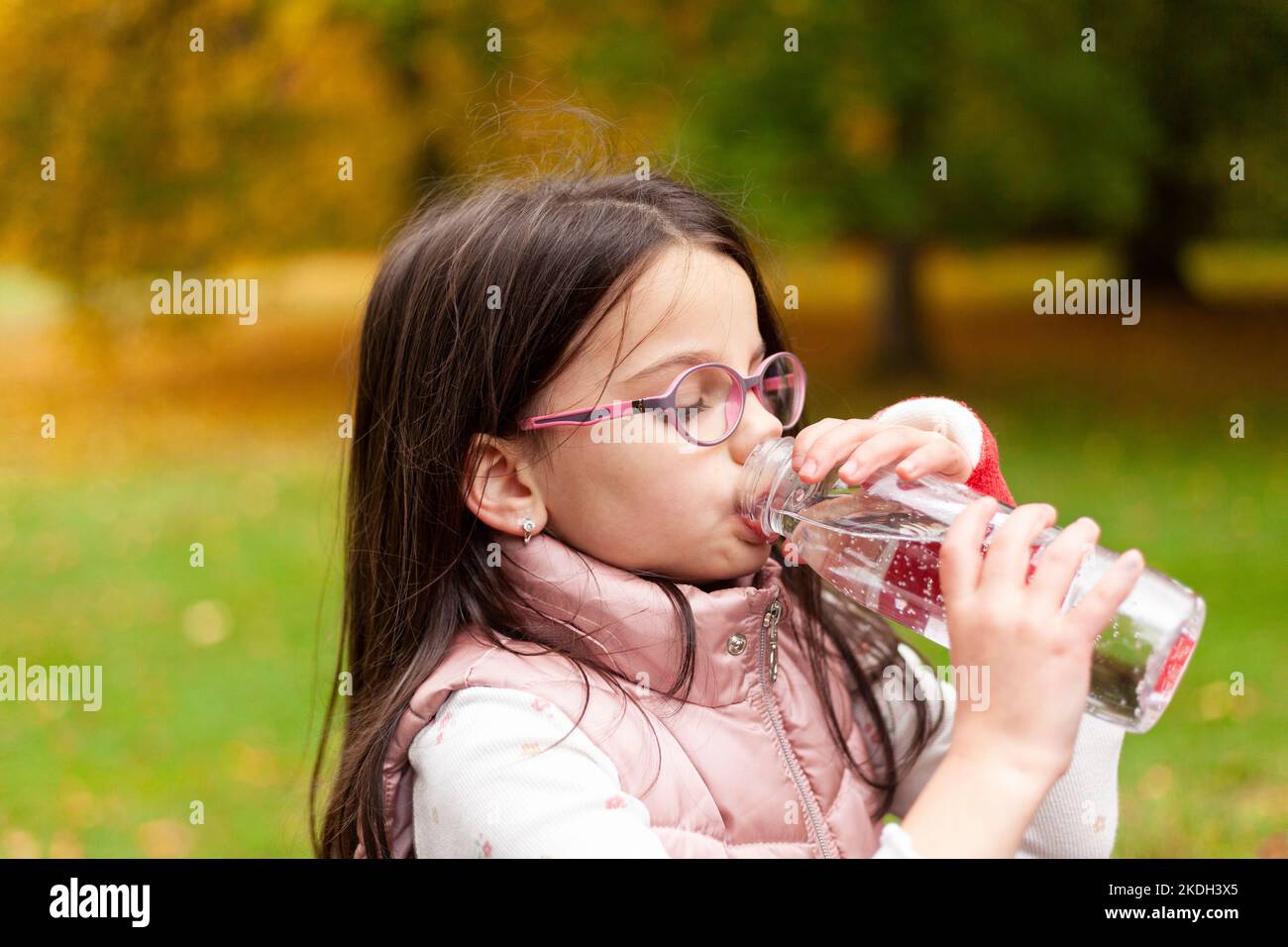 Cute girl with long hair and glasses drinks water from a plastic bottle on a blurred autumn park background Stock Photo