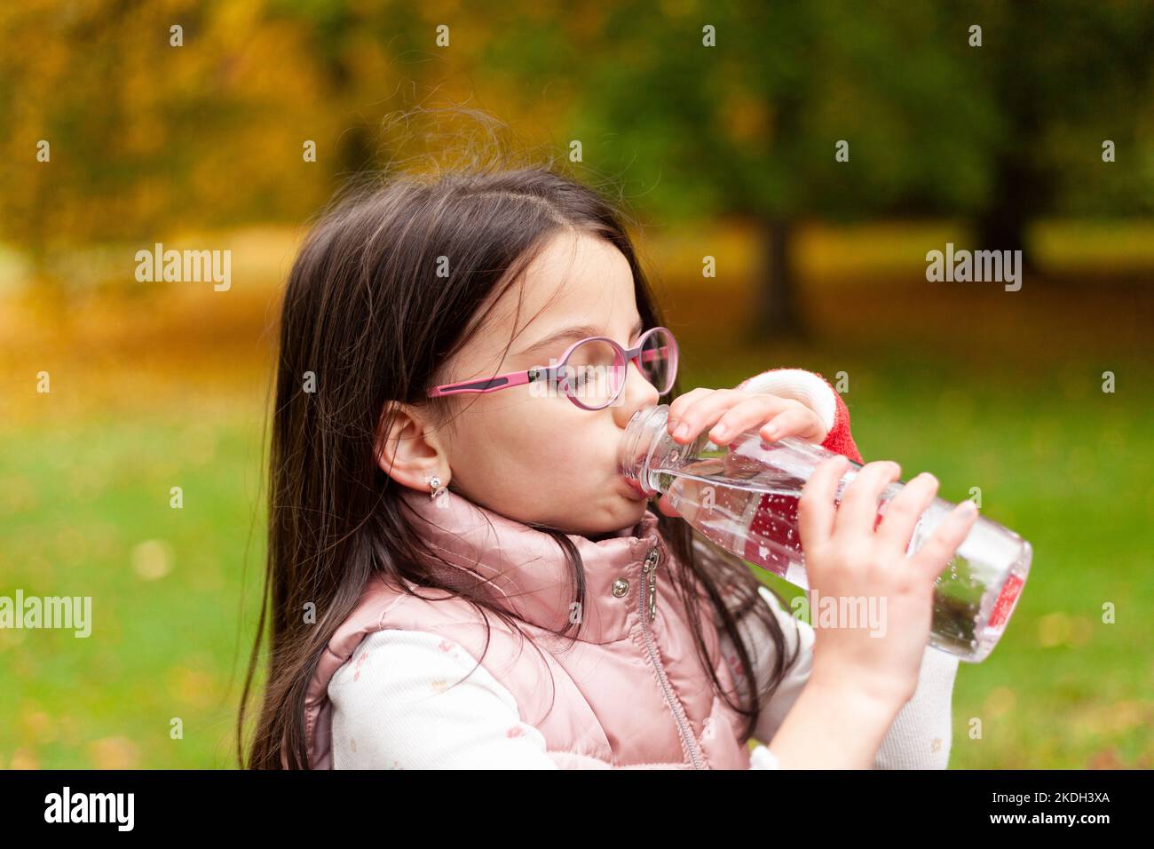 Cute girl with long hair and glasses drinks water from a plastic bottle on a blurred autumn park background Stock Photo
