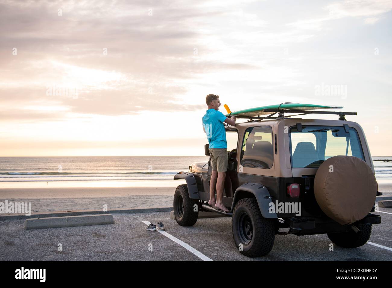 Young Man with surfboard on top of car at sunrise at Beach Stock Photo