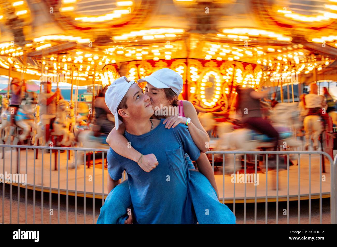 Teen girl kissing cheek of shocked teen boy in front of carousel Stock Photo