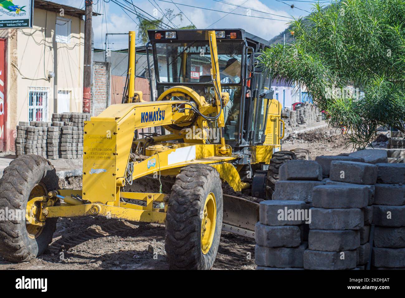 Motor grader working on finishing work on sewer system pipes below a street paved in concrete cast blocks. Stock Photo