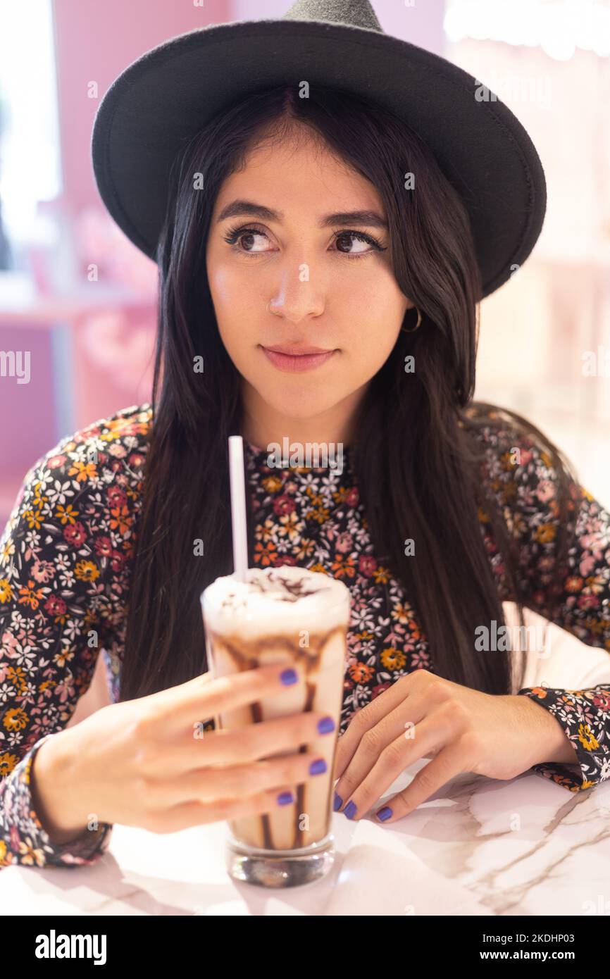 young woman wearing hat and patterned dress holding glass of cold chocolate drink as sweet, latin model lifestyle Stock Photo