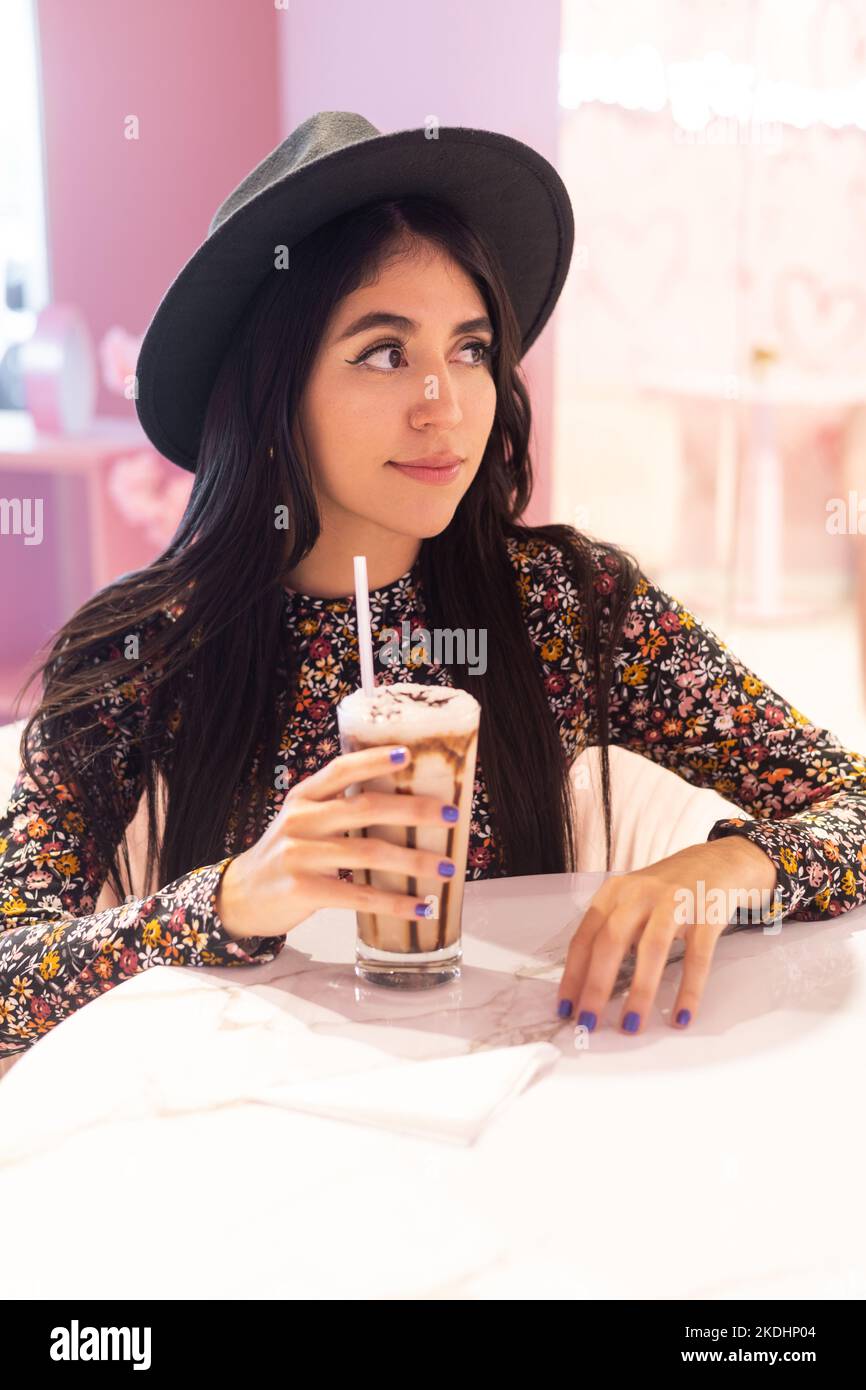 sitting young woman wearing hat and patterned dress with glass of cold chocolate drink with sorbet, latin model lifestyle, beauty Stock Photo